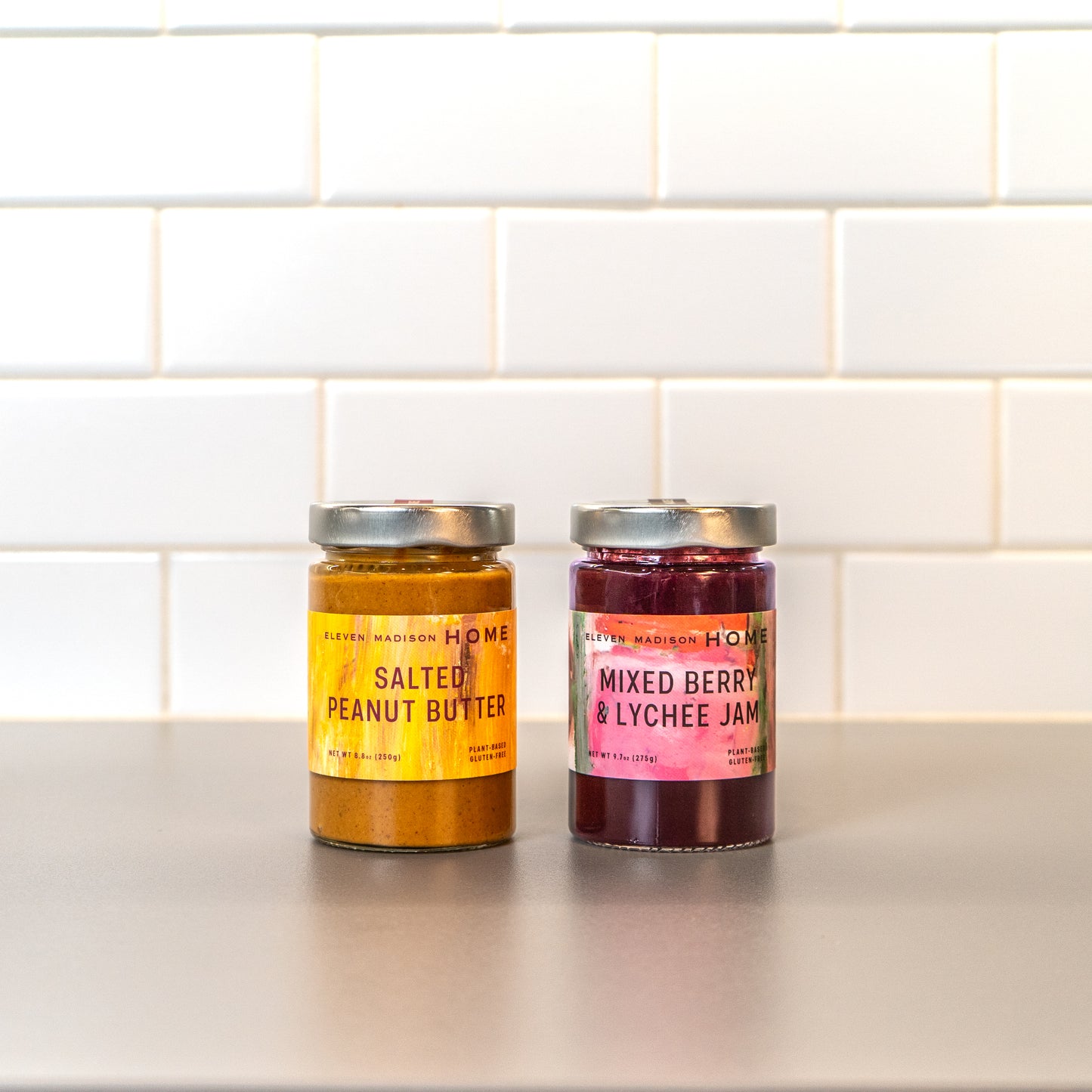 Jar of Salted Peanut Butter beside a jar of Mixed Berry & Lychee Jam on a kitchen counter with a white tiled background. 